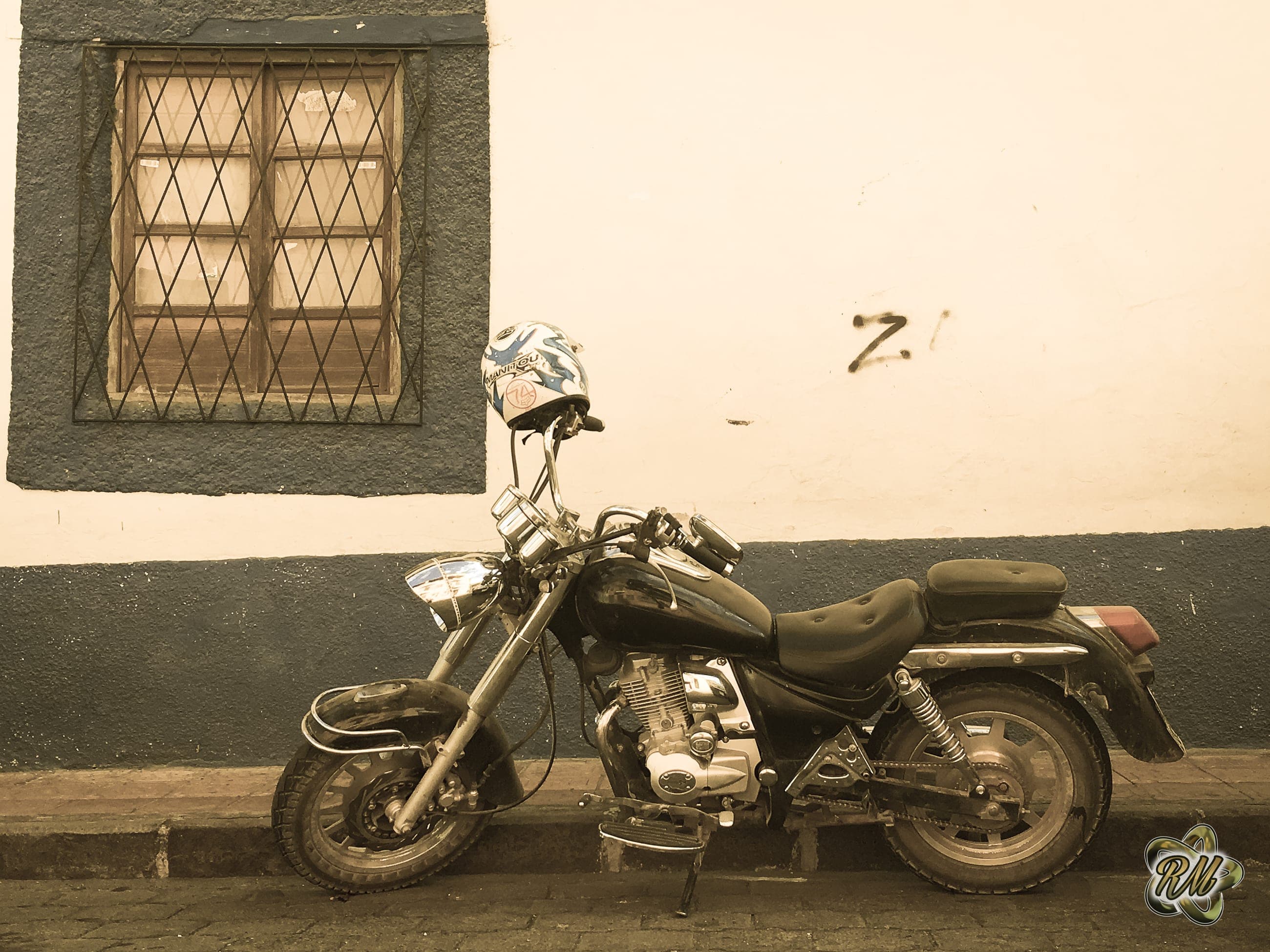 Sepia-toned cruiser motorcycle parked against a weathered wall with a barred window